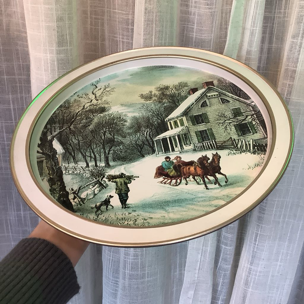 Decorative plate with a winter scene of a house and people on horseback, held against a curtain background.