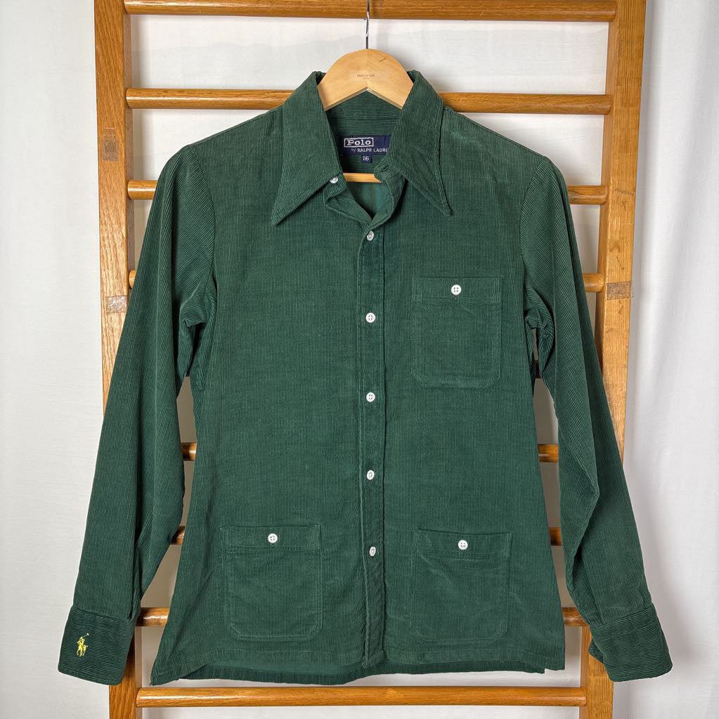 A green corduroy button-up long sleeve shirt on a wooden rack against a white wall.