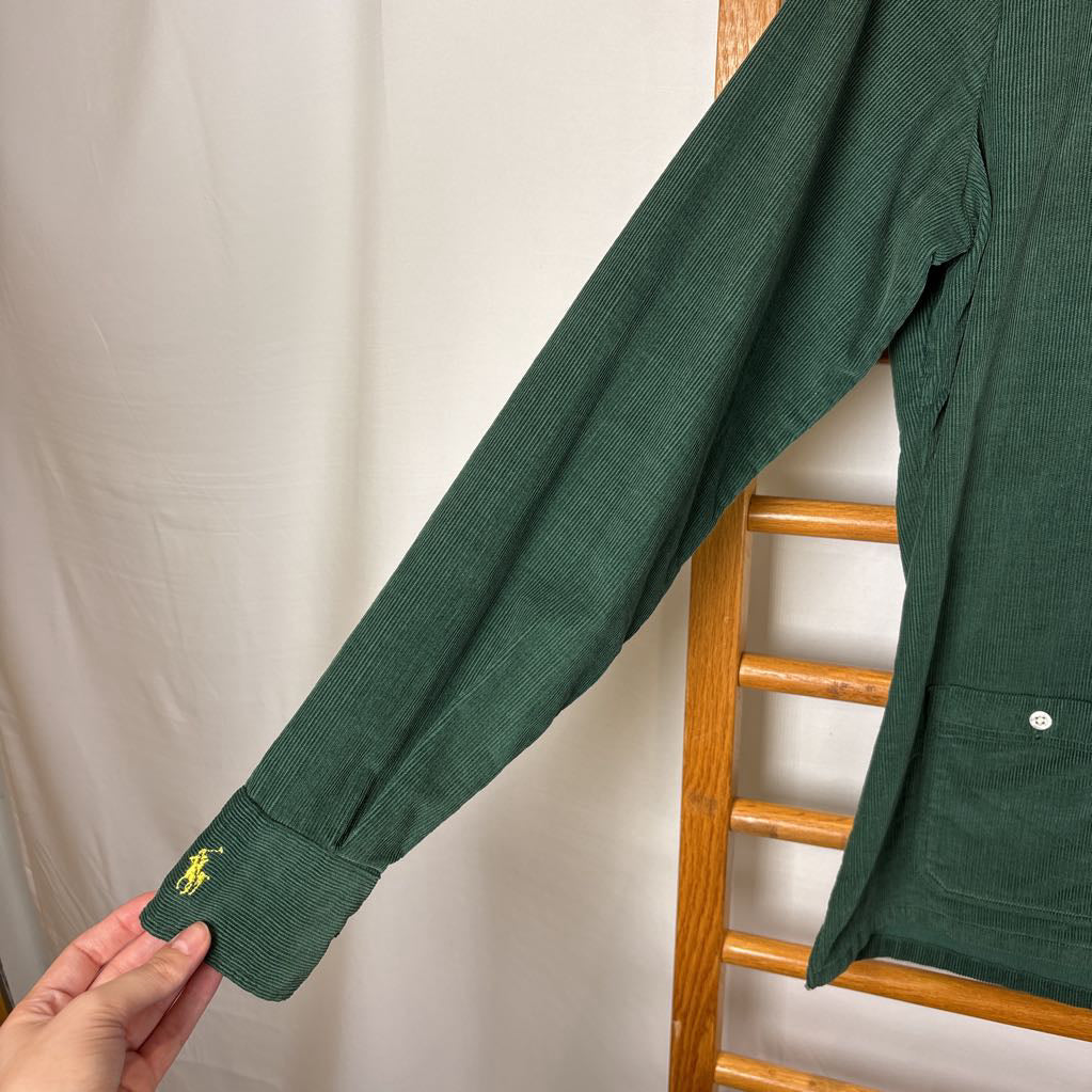 A green corduroy button-up long sleeve shirt on a wooden rack against a white wall.