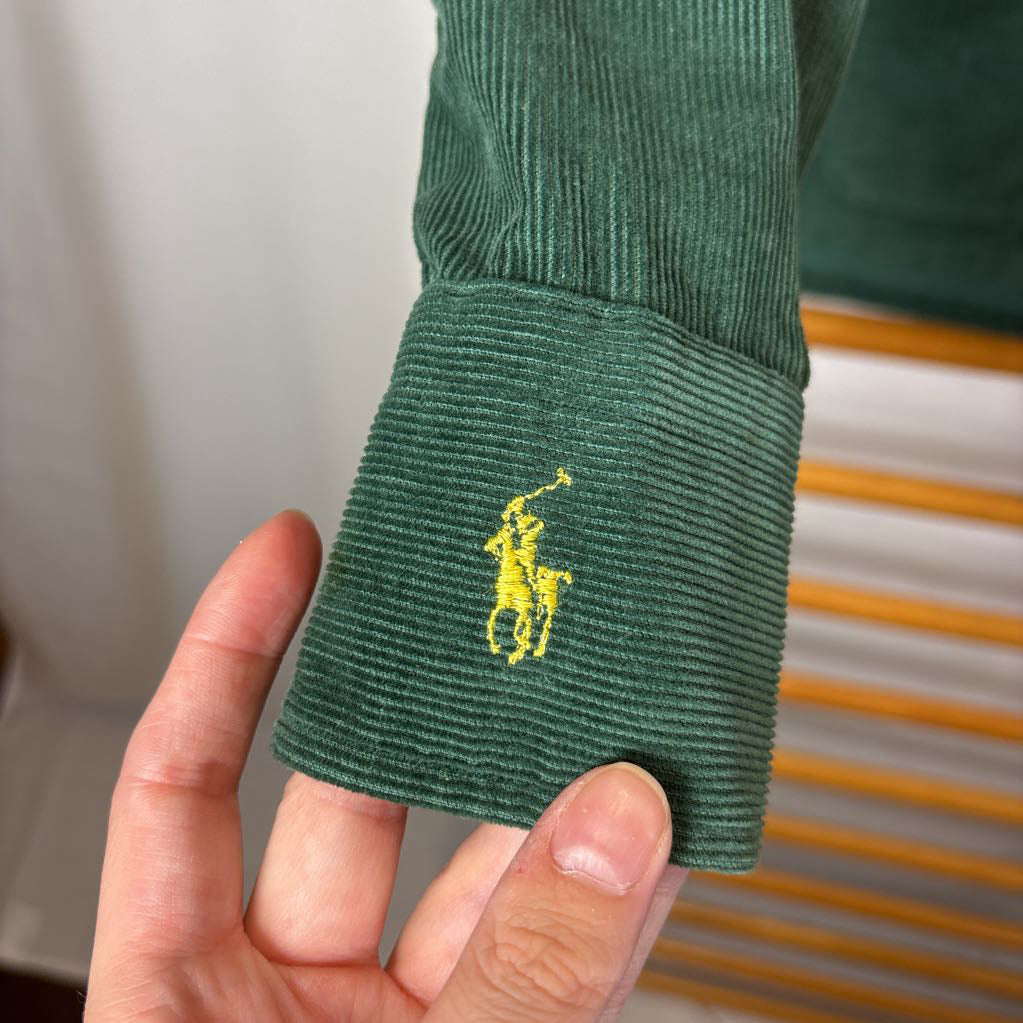 A green corduroy button-up long sleeve shirt on a wooden rack against a white wall.