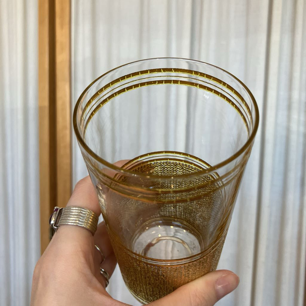 Hand holding a gold embossed glass tilting it towards the camera up against a white fabric background