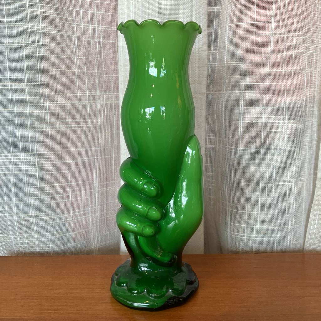 Green glass vase shaped like a hand holding a vase against a textured white curtain background.