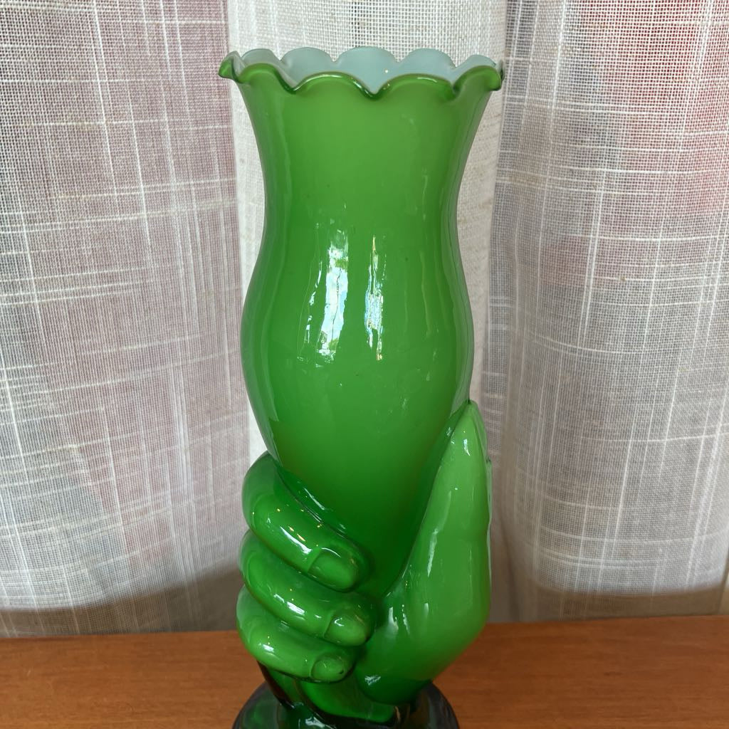Green glass vase shaped like a hand holding a vase against a textured white curtain background.