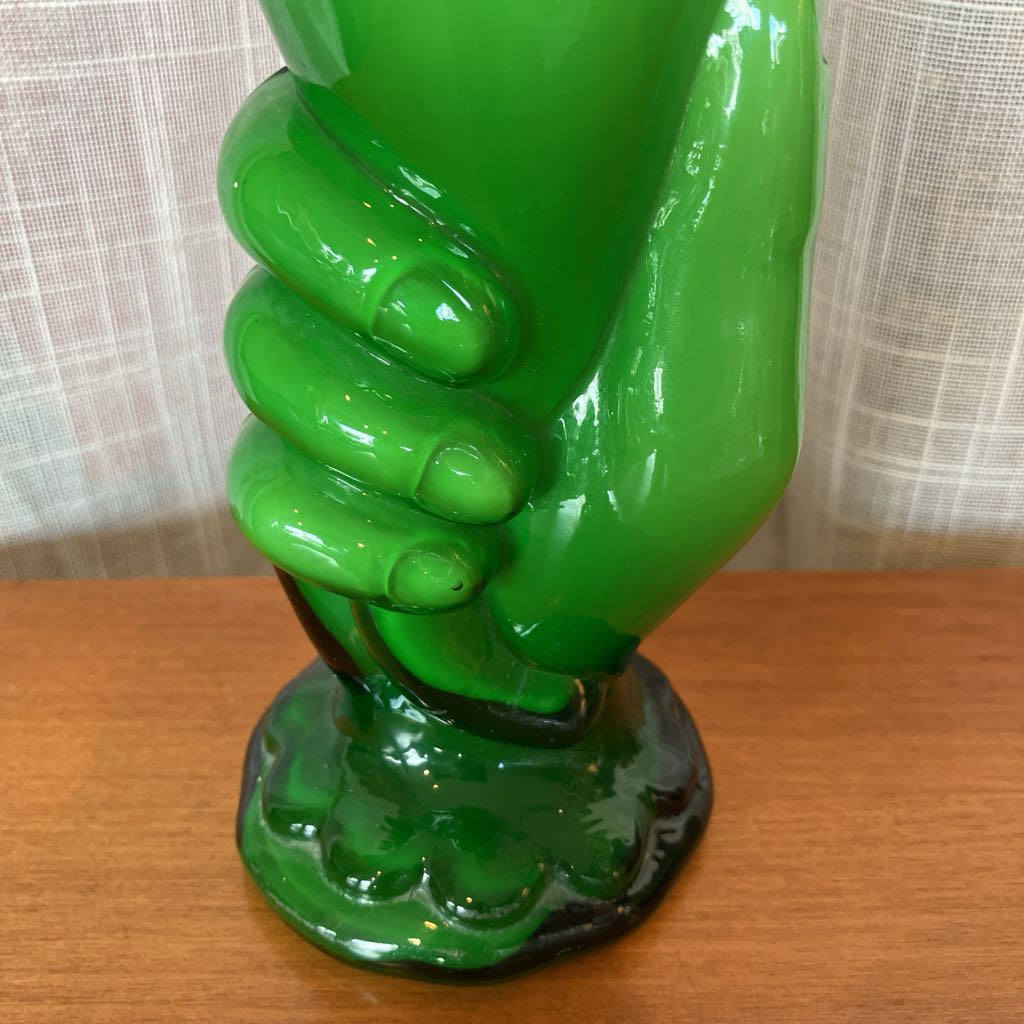 Green glass vase shaped like a hand holding a vase against a textured white curtain background.