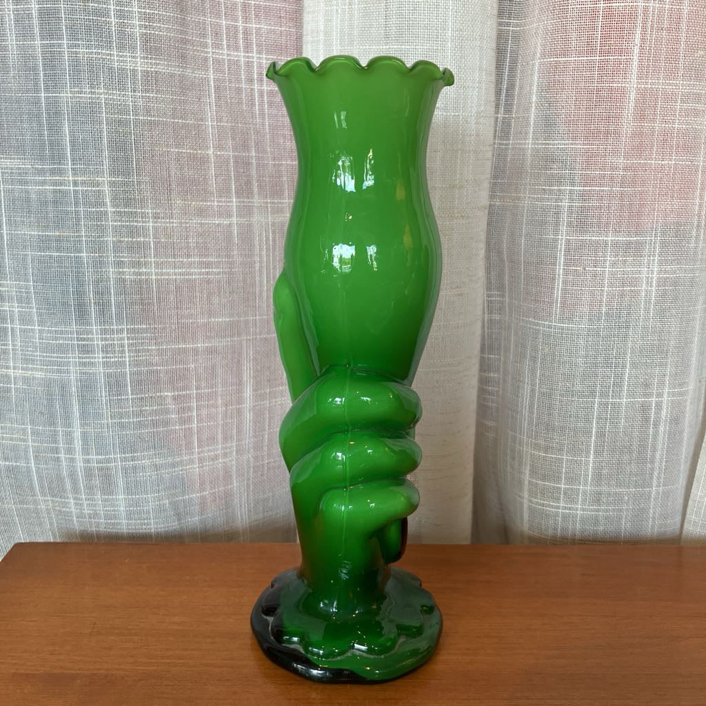 Green glass vase shaped like a hand holding a vase against a textured white curtain background.