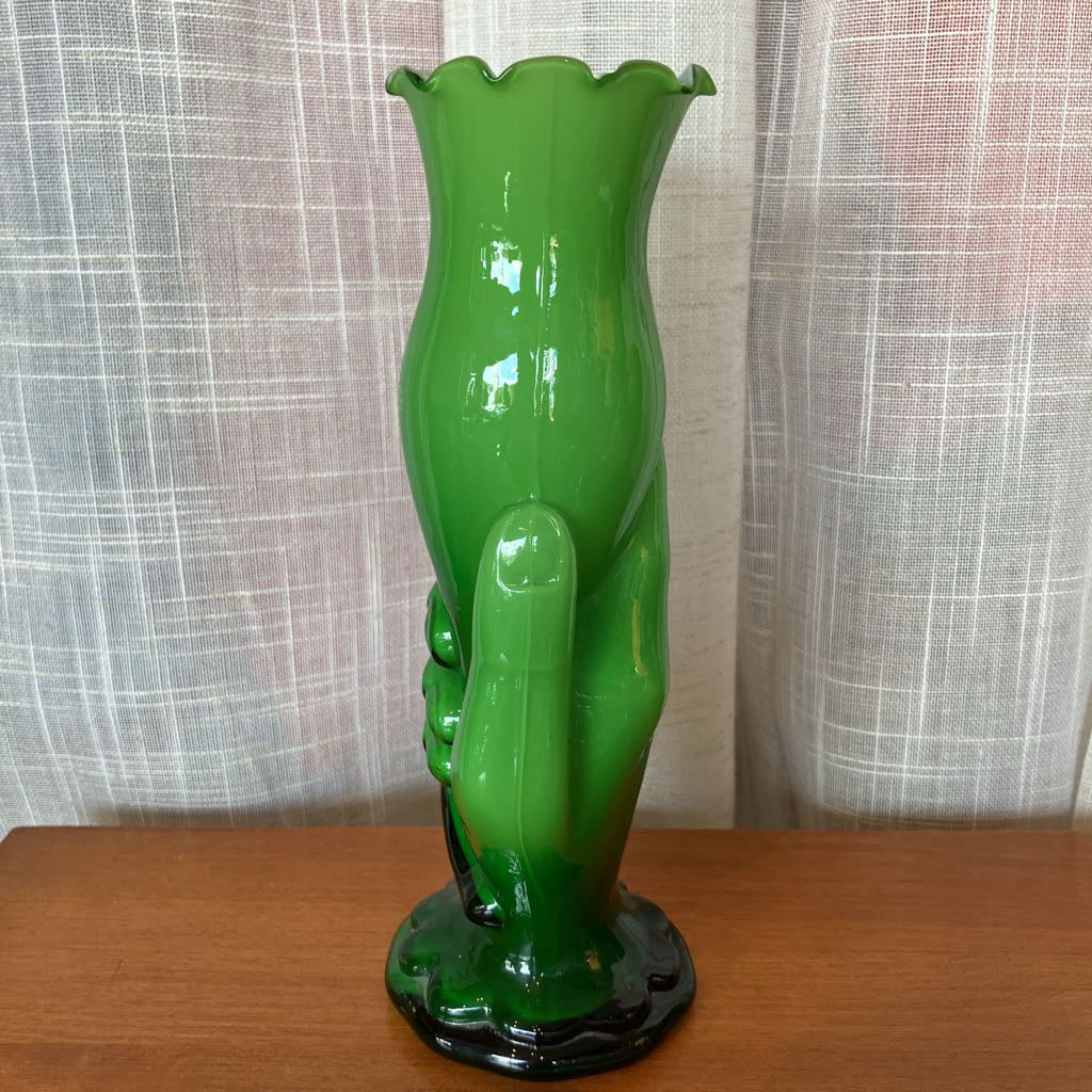 Green glass vase shaped like a hand holding a vase against a textured white curtain background.