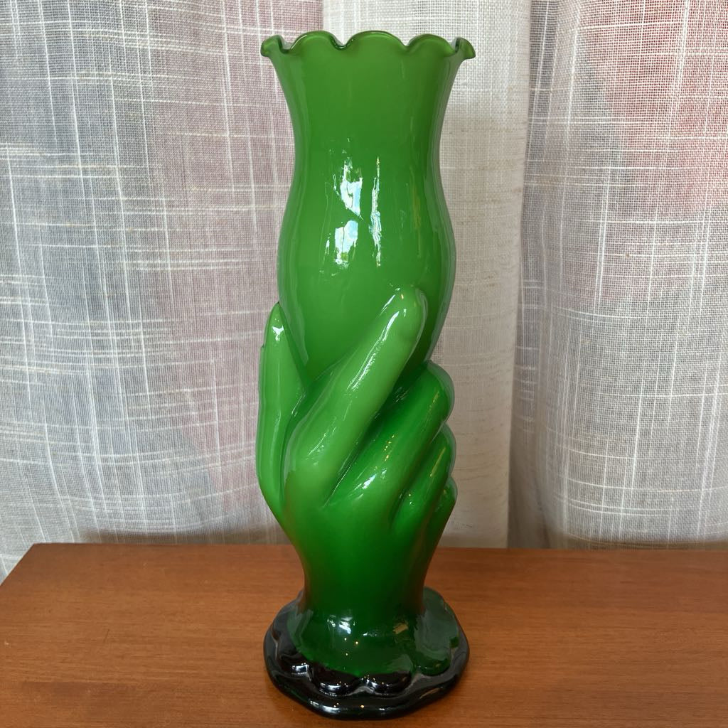 Green glass vase shaped like a hand holding a vase against a textured white curtain background.
