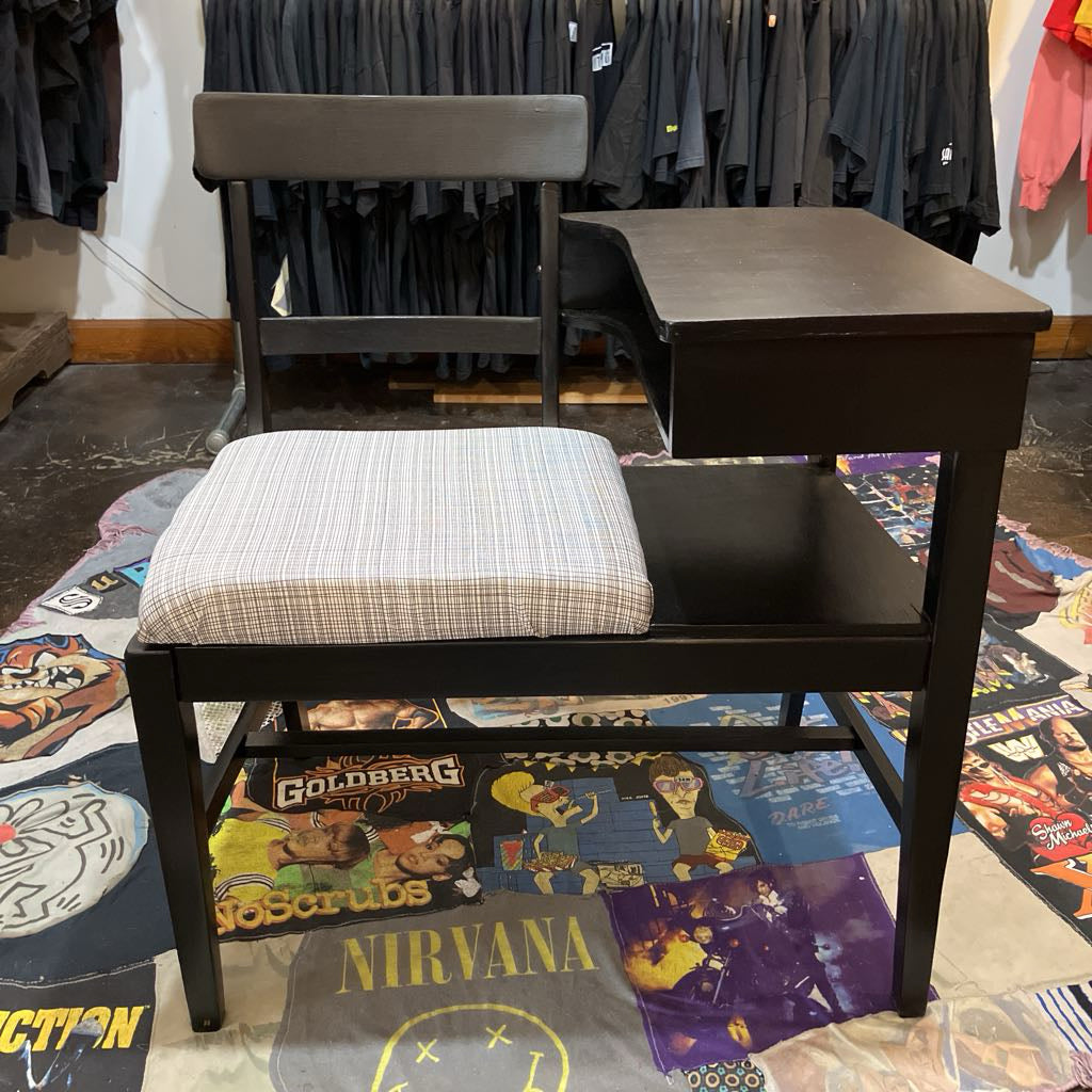 Black desk with cushioned chair on a colorful floor with t-shirts hanging in the background