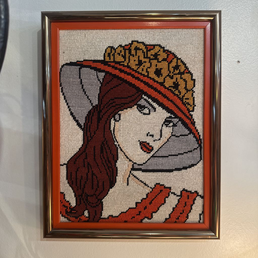 Framed cross-stitched artwork of a woman wearing a hat with a leopard print design.