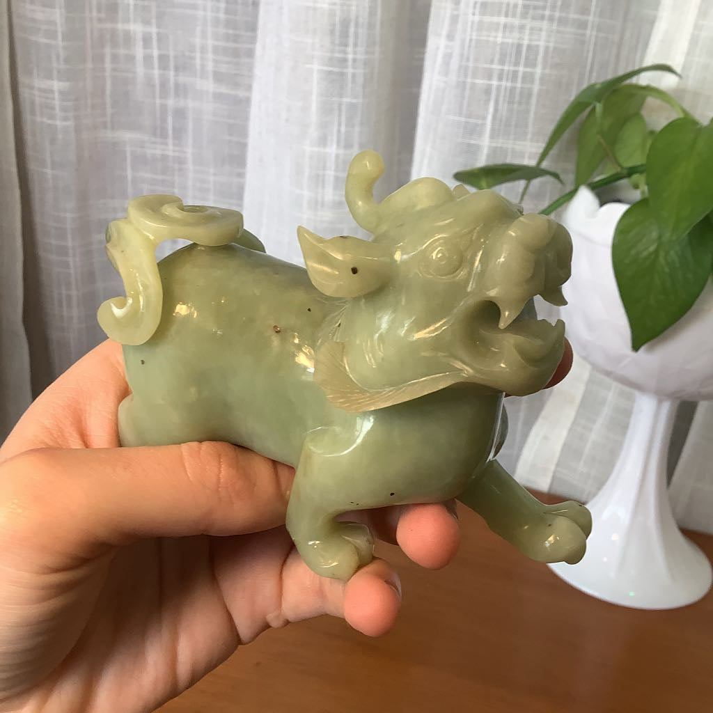 Hand holding a green ceramic dragon figurine with a plant and white curtain in the background