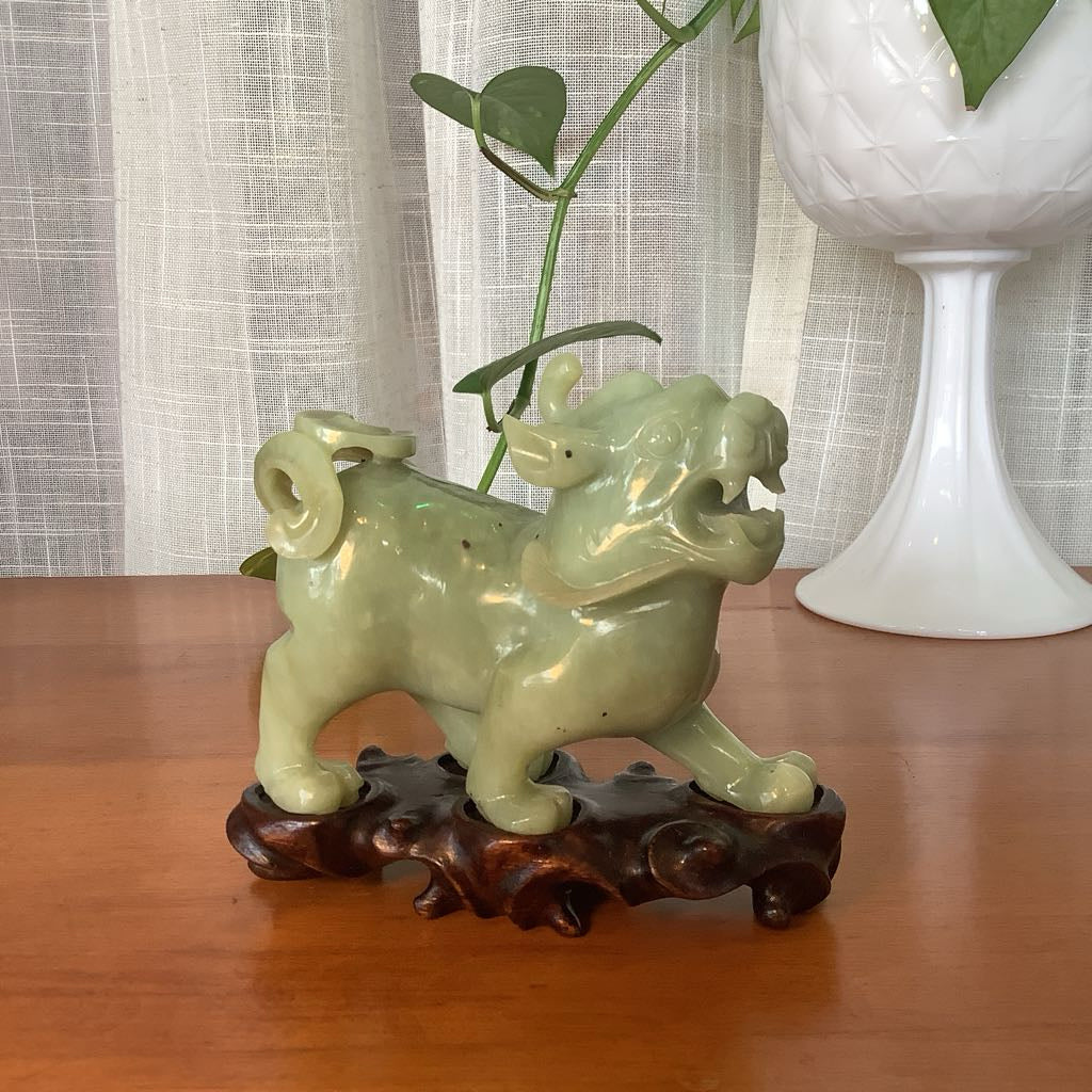 Green ceramic dog figurine on a wooden stand with a white vase and plant in the background.