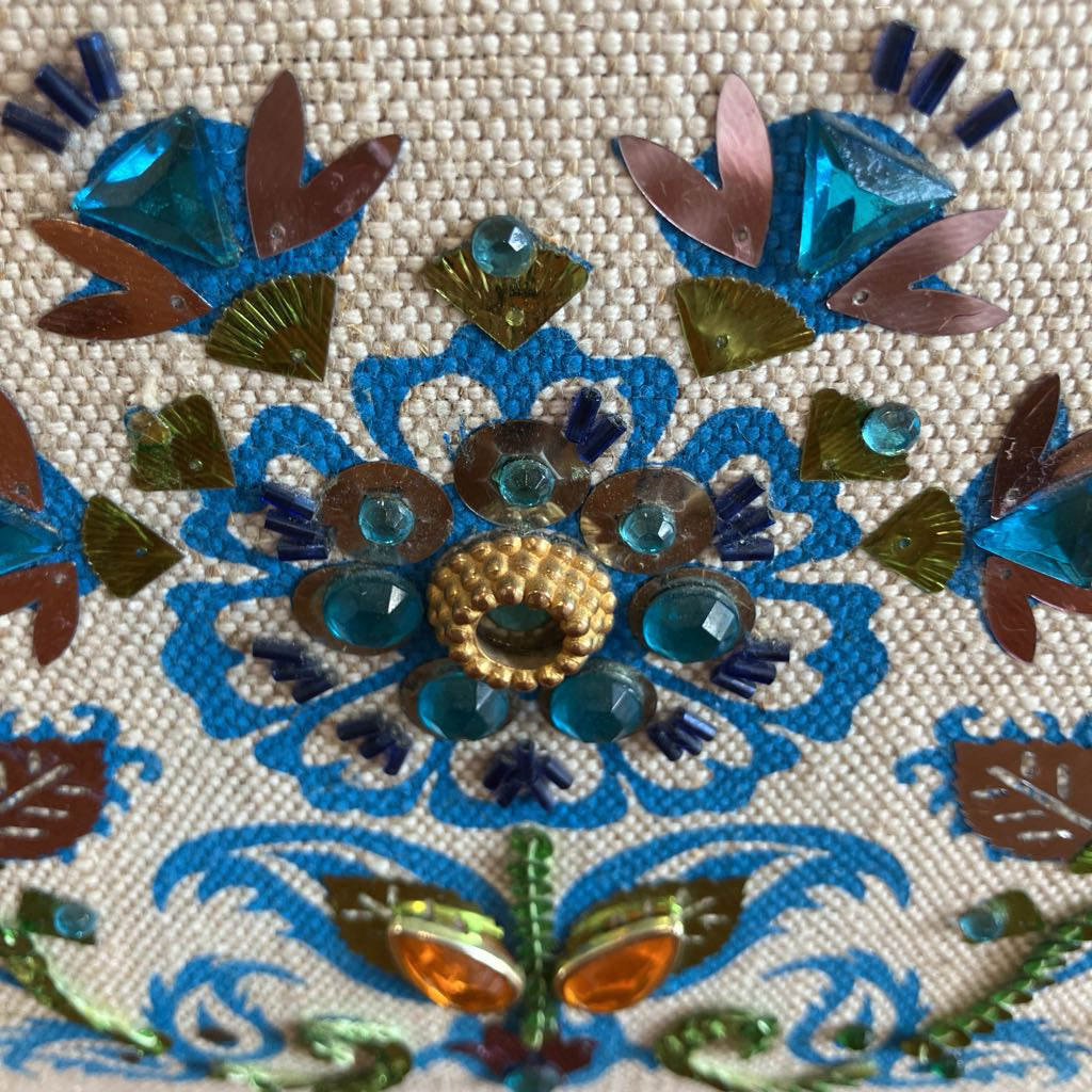 Close-up of decorative embroidery with blue, green, and gold colors on a beige fabric background.