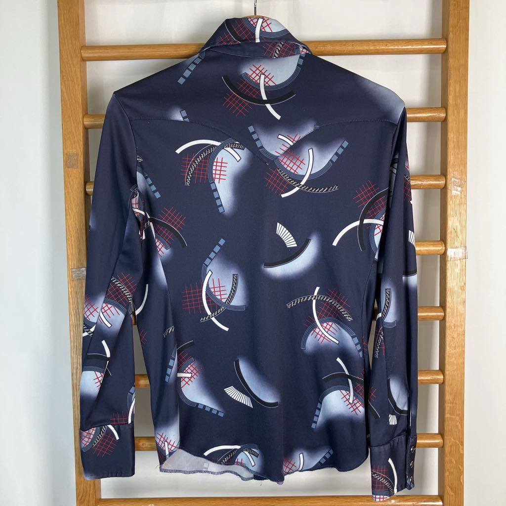 A long-sleeve button-up shirt in navy blue with a graphic print of baseball caps, photographed hanging on a wooden hanger rack against a light wall.