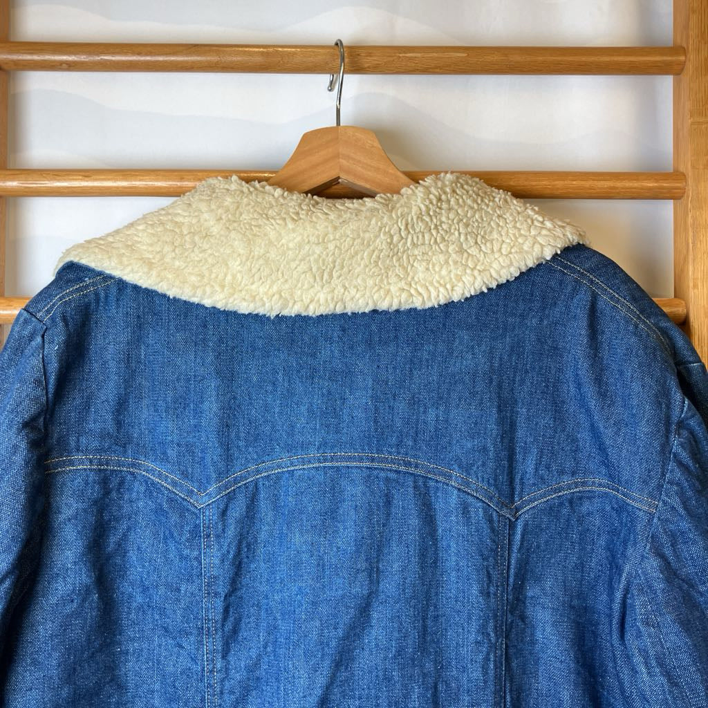 Blue denim jacket with cream collar on a wooden hanger against a white wall.