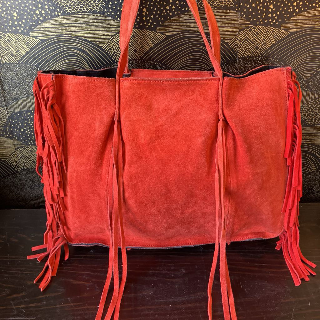 Red suede handbag with fringe details on a wooden surface with a patterned background