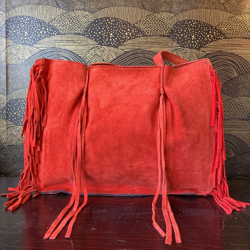 Red suede handbag with fringe details on a wooden surface against a patterned wall.