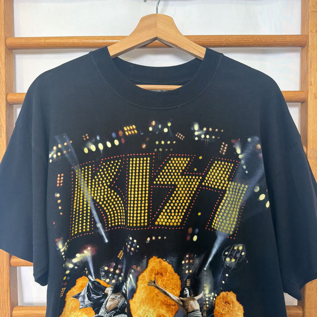 A black short-sleeve graphic t-shirt hanging on a wooden hanger against a wooden window frame. The shirt features a large yellow 'KISS' logo made of dots, with a concert scene graphic below.