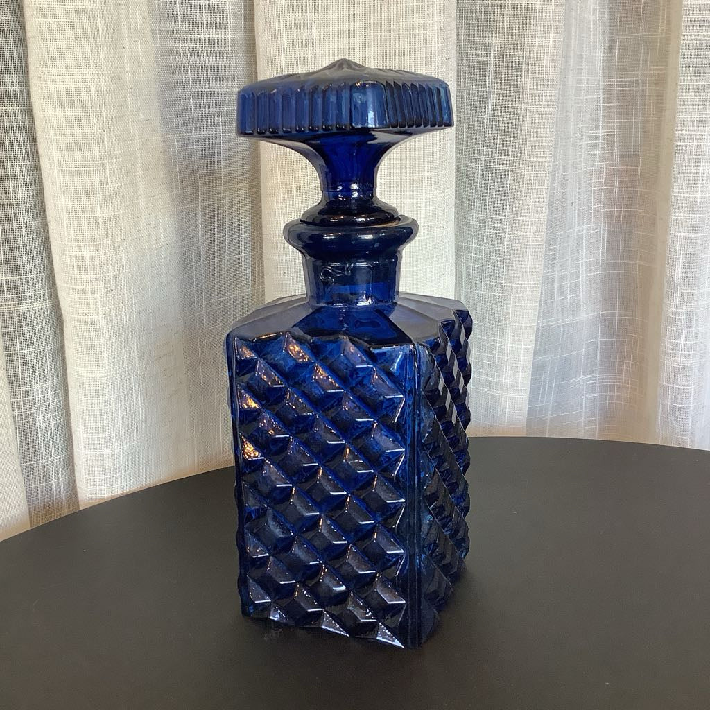 Blue glass decanter with textured design on a dark surface and light curtain background