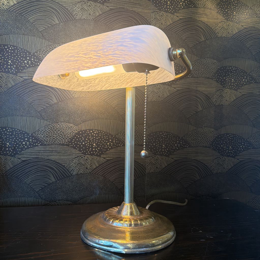 Vintage-style desk lamp with a textured shade on a wooden surface against a patterned wallpaper background.