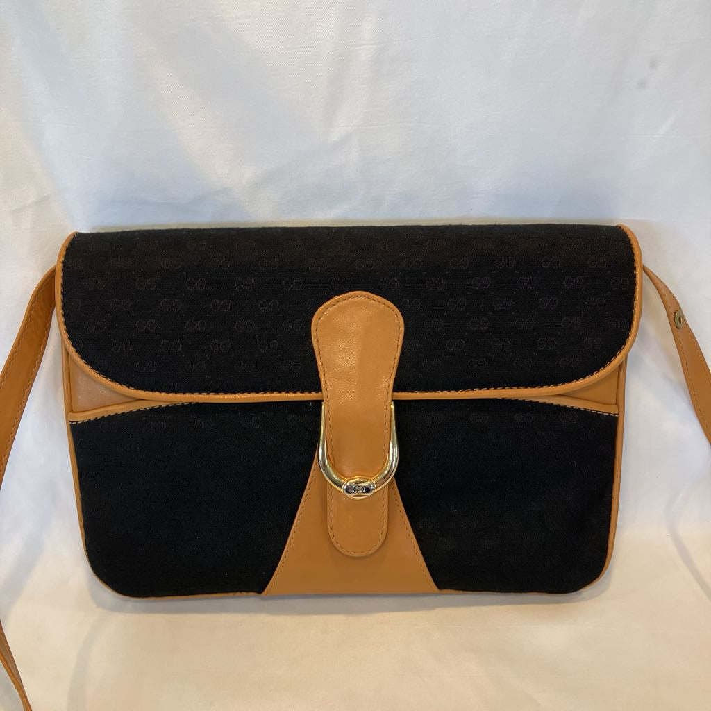 A black suede shoulder bag with tan leather trim and a central strap closure, displayed against a white fabric backdrop.