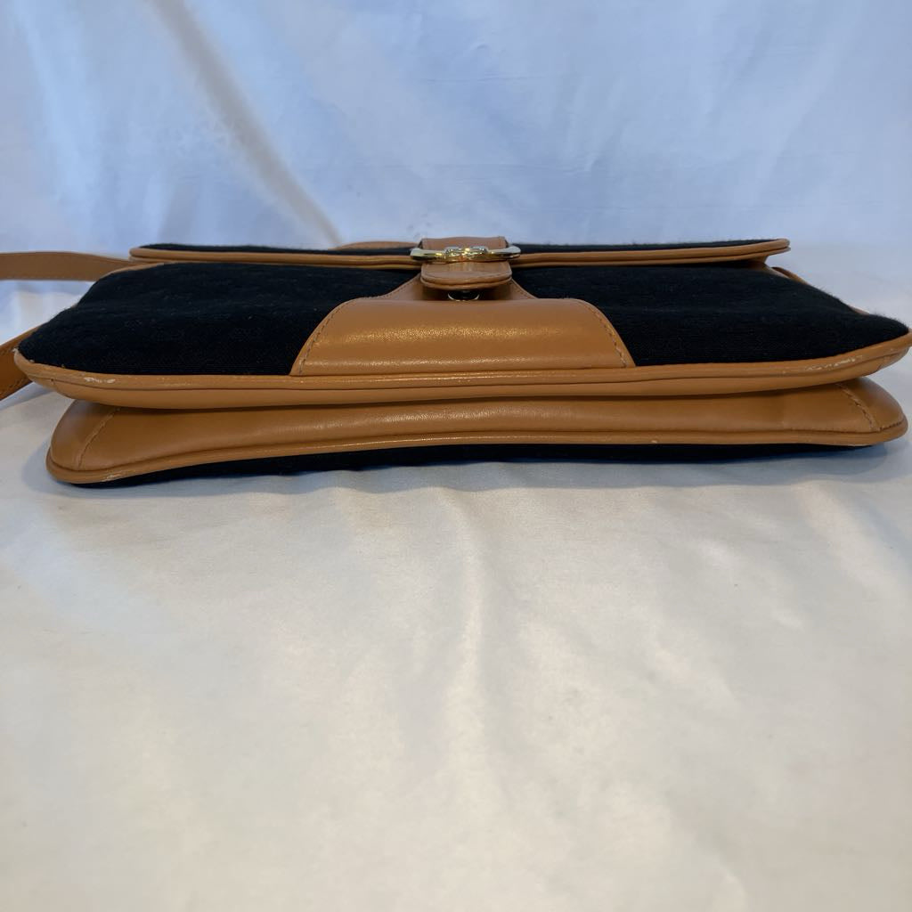A vintage shoulder bag with a black suede body and tan leather trim, featuring a central gold-tone metal clasp and a thin strap extending to the left, displayed on a white fabric background.