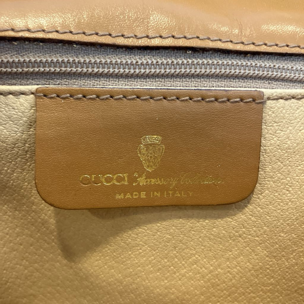 Close-up of a Gucci leather bag showing a tan leather patch with gold embossed logo and 'GUCCI Missory trinkets MADE IN ITALY' text, with a gray zipper above.