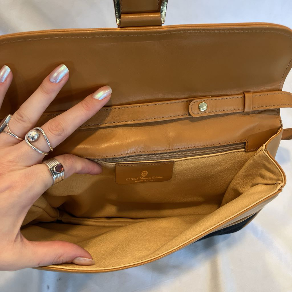 A tan leather handbag shown open, with a hand holding the interior flap. The bag has a matching tan interior lining, a zip pocket, and a small gold-tone logo plaque.