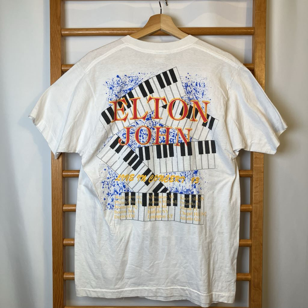 White t-shirt with Elton John graphic design on a wooden hanger against a white wall.