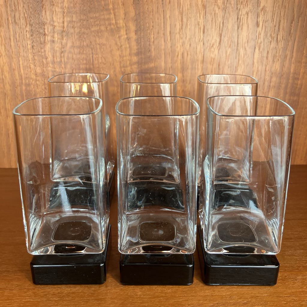 Set of six clear glasses with black bases on a wooden surface.