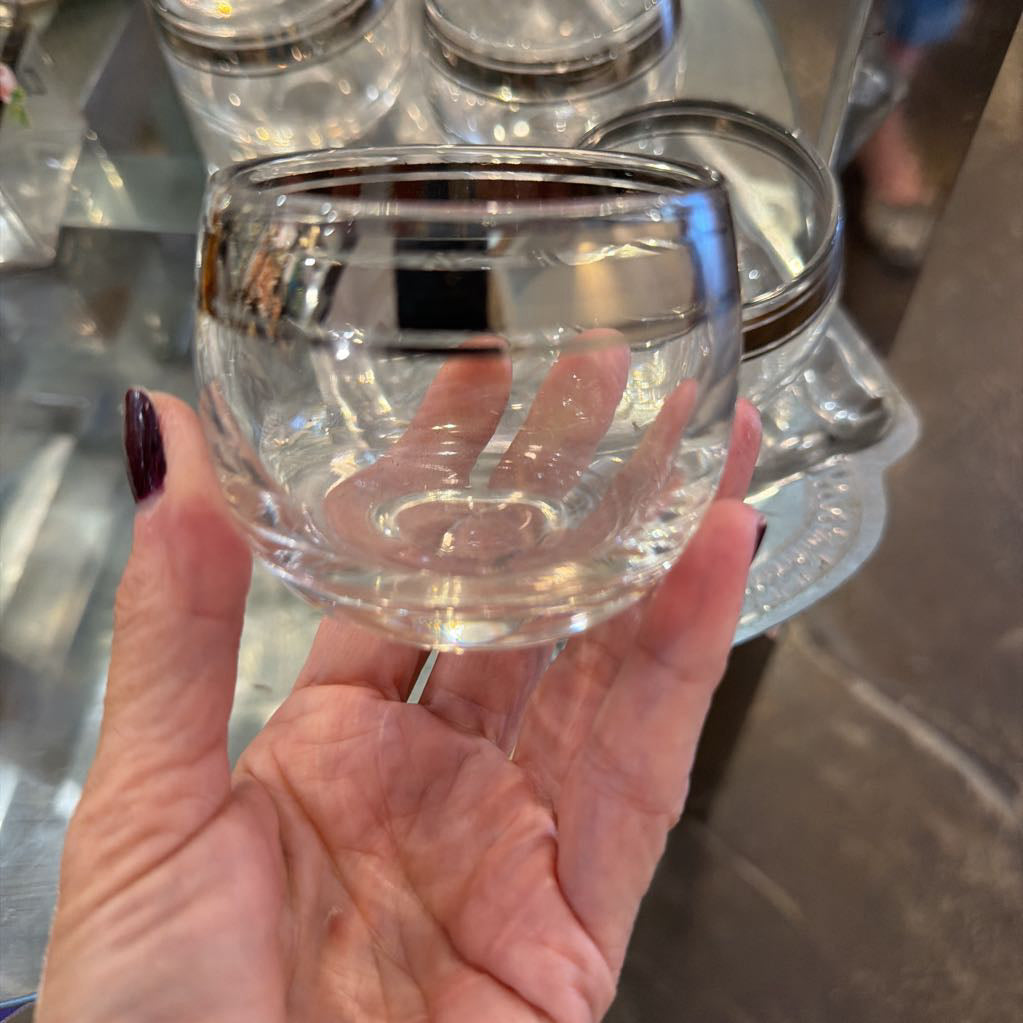 A hand with dark purple nail polish holds a small, clear glass bowl or ramekin in front of a glass display shelf.