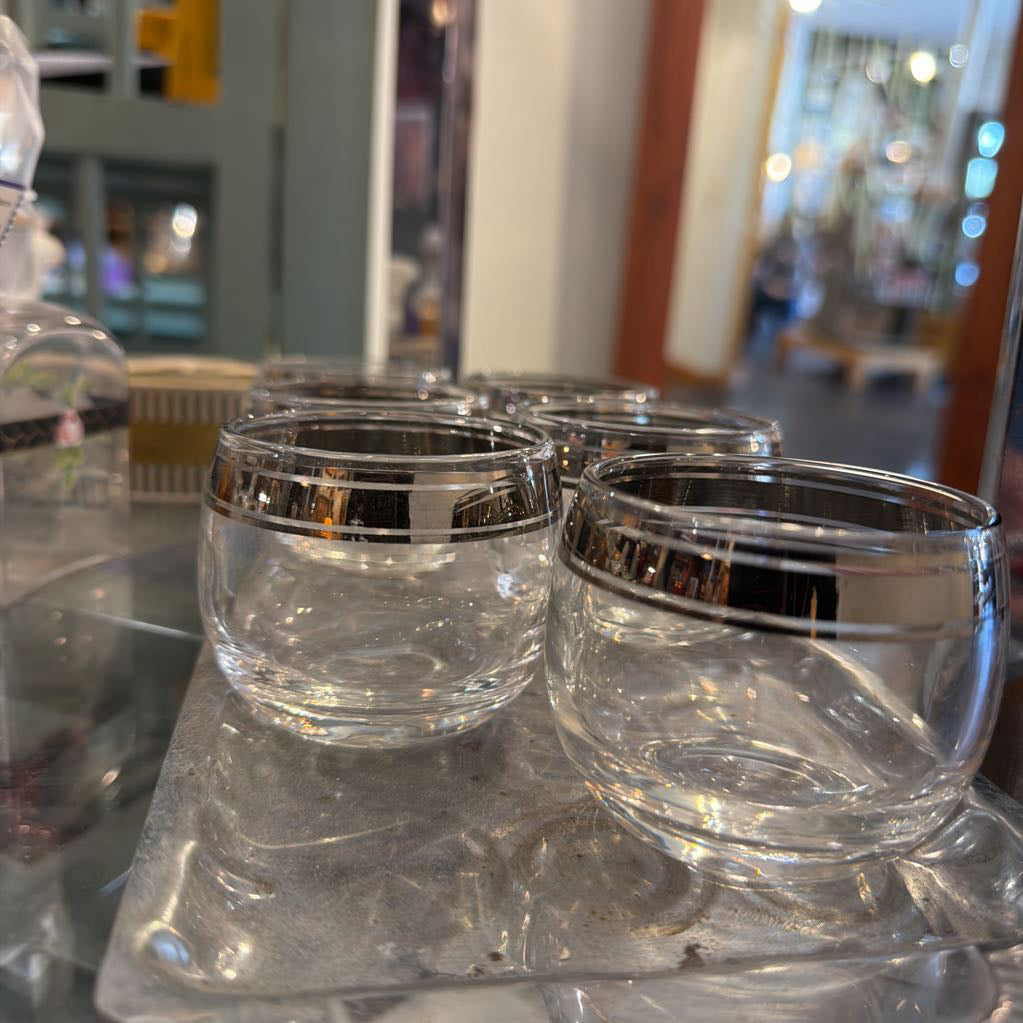 A set of clear glass cups with a shiny silver metallic band around the rim, displayed on a glass surface with a blurred shop background.