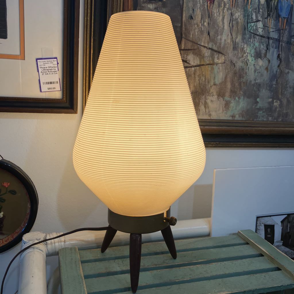 Table lamp with a textured shade on a wooden surface, with artworks in the background.
