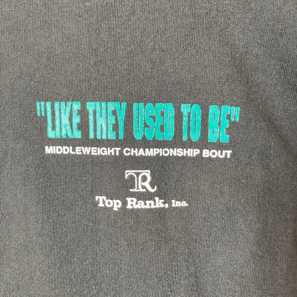 Close-up of a dark gray heathered fabric surface with printed text reading "LIKE THEY USED TO BE" in bright turquoise, followed by "MIDDLEWEIGHT CHAMPIONSHIP BOUT" and a white Top Rank, Inc. logo.
