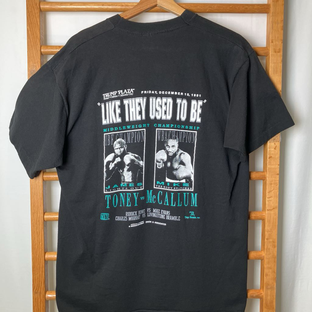 A black graphic t-shirt hanging on a wooden hanger against a wooden chair, showing the back of the shirt with a vintage boxing event print featuring Toney McCallum.