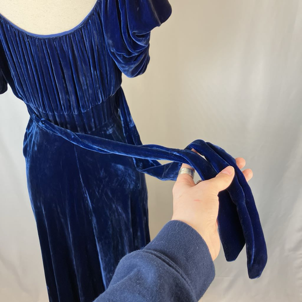 Blue velvet dress with a hand adjusting the belt against a neutral background