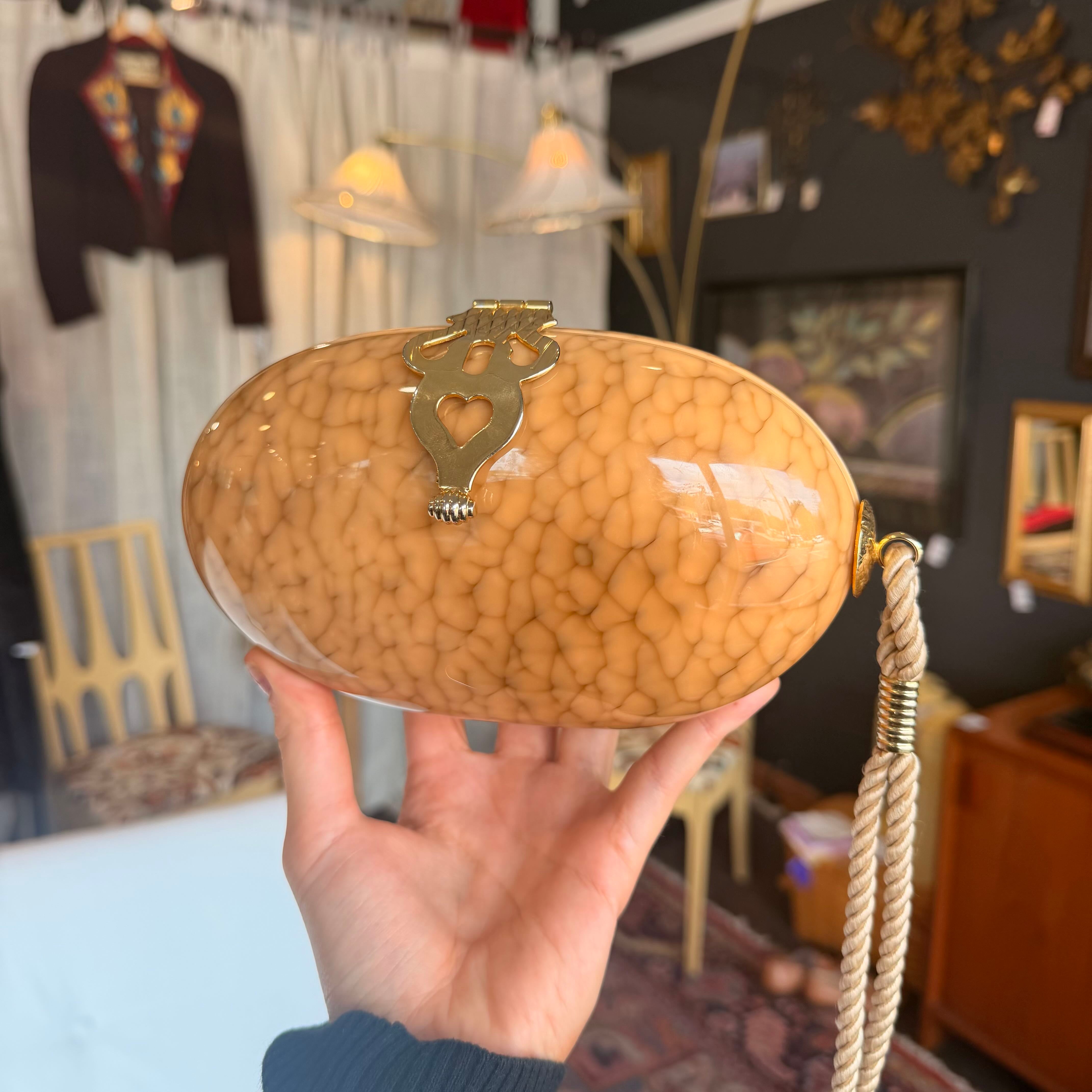 Hand holding a textured beige clutch bag with a decorative clasp in an indoor setting.
