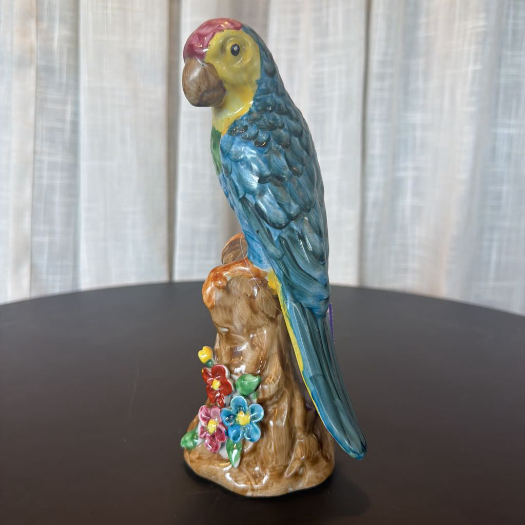 Colorful ceramic parrot figurine on a wooden base with flowers against a white curtain background