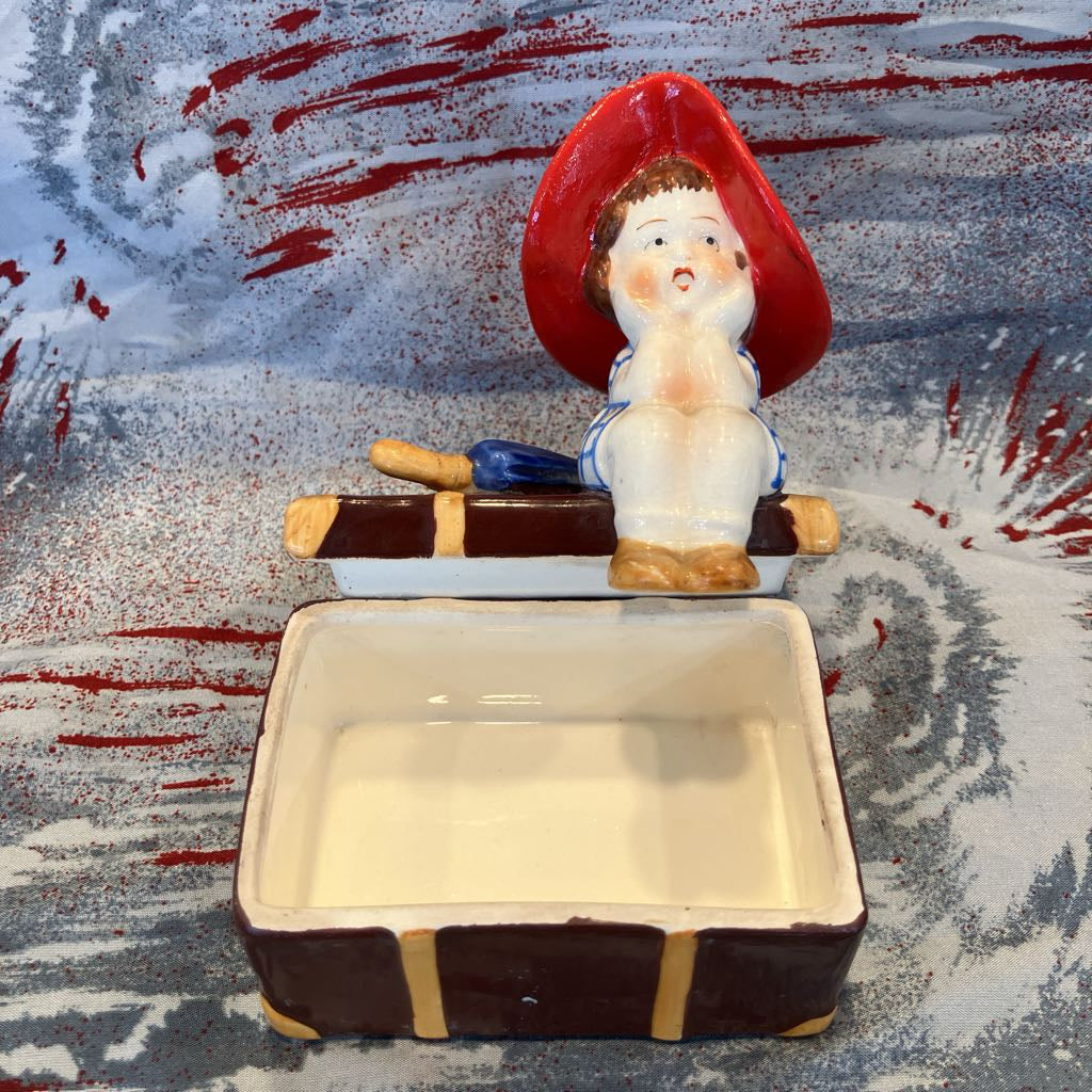 Ceramic figure of a girl with a red hat on top of a rectangular dish with a textured background.