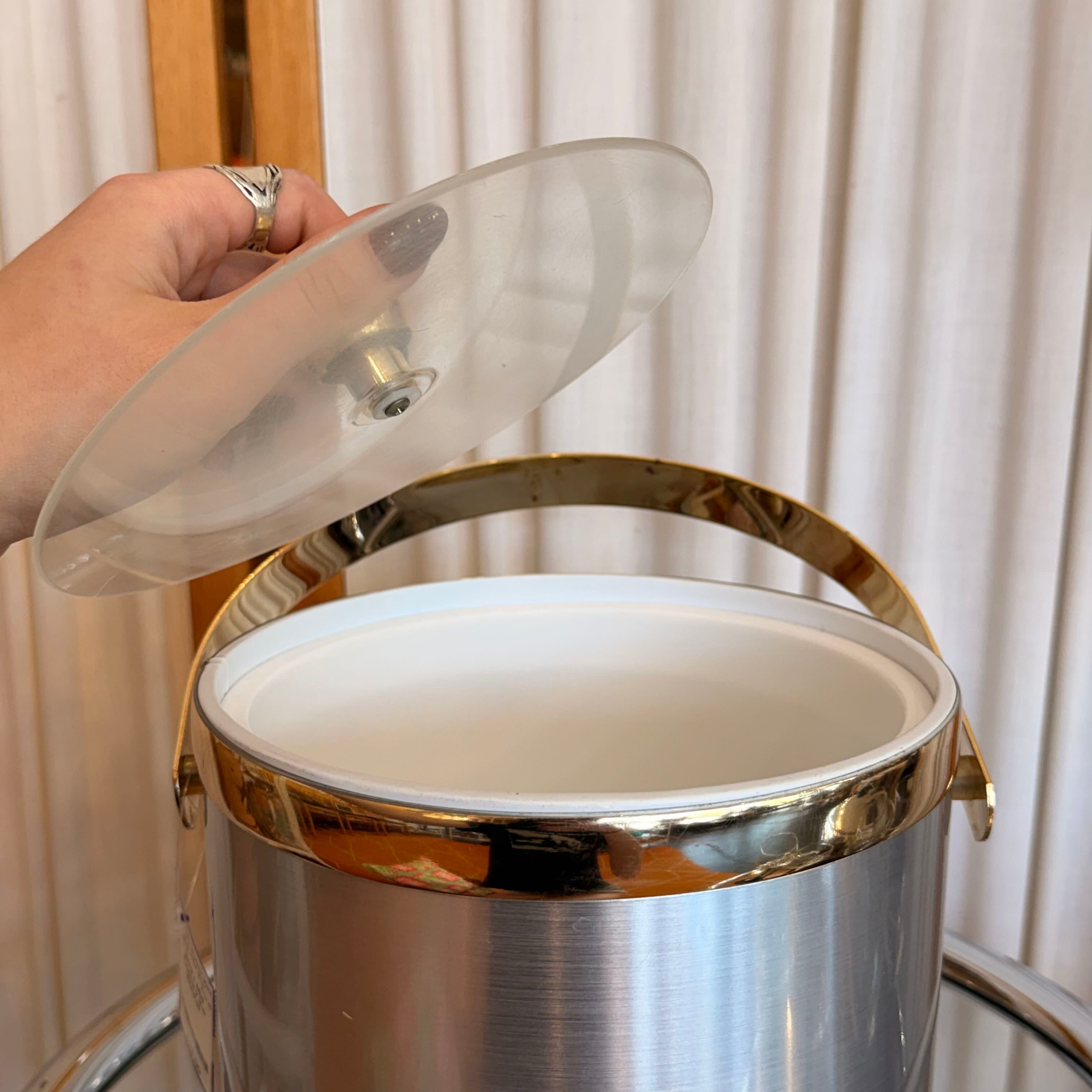 Stainless steel container with a clear lid being held by a hand against a white curtain background