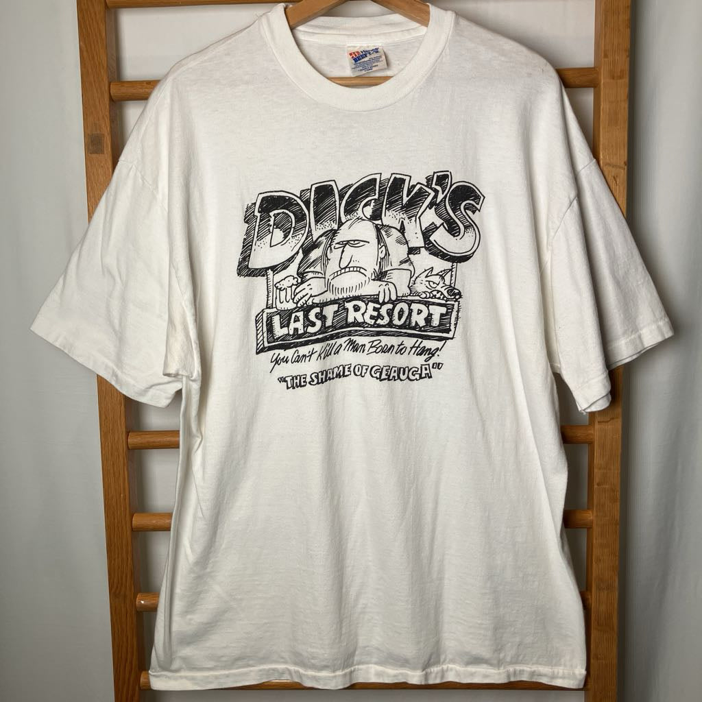 A white vintage graphic t-shirt hanging on a wooden hanger against a wooden chair. The graphic features a black-and-white illustration of a bearded man with a pipe and the text 'Dicky's Last Resort'.