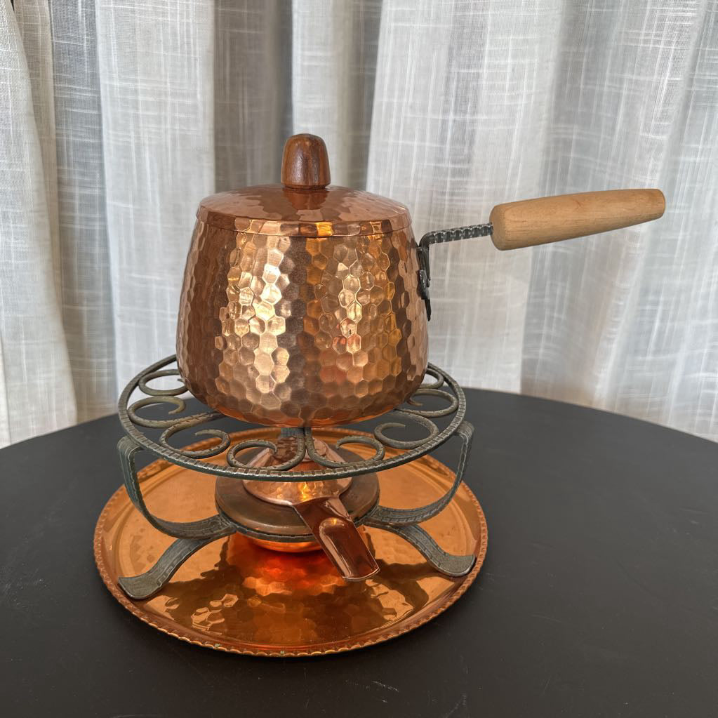 Copper fondue pot with wooden handle on a black surface and white curtain background