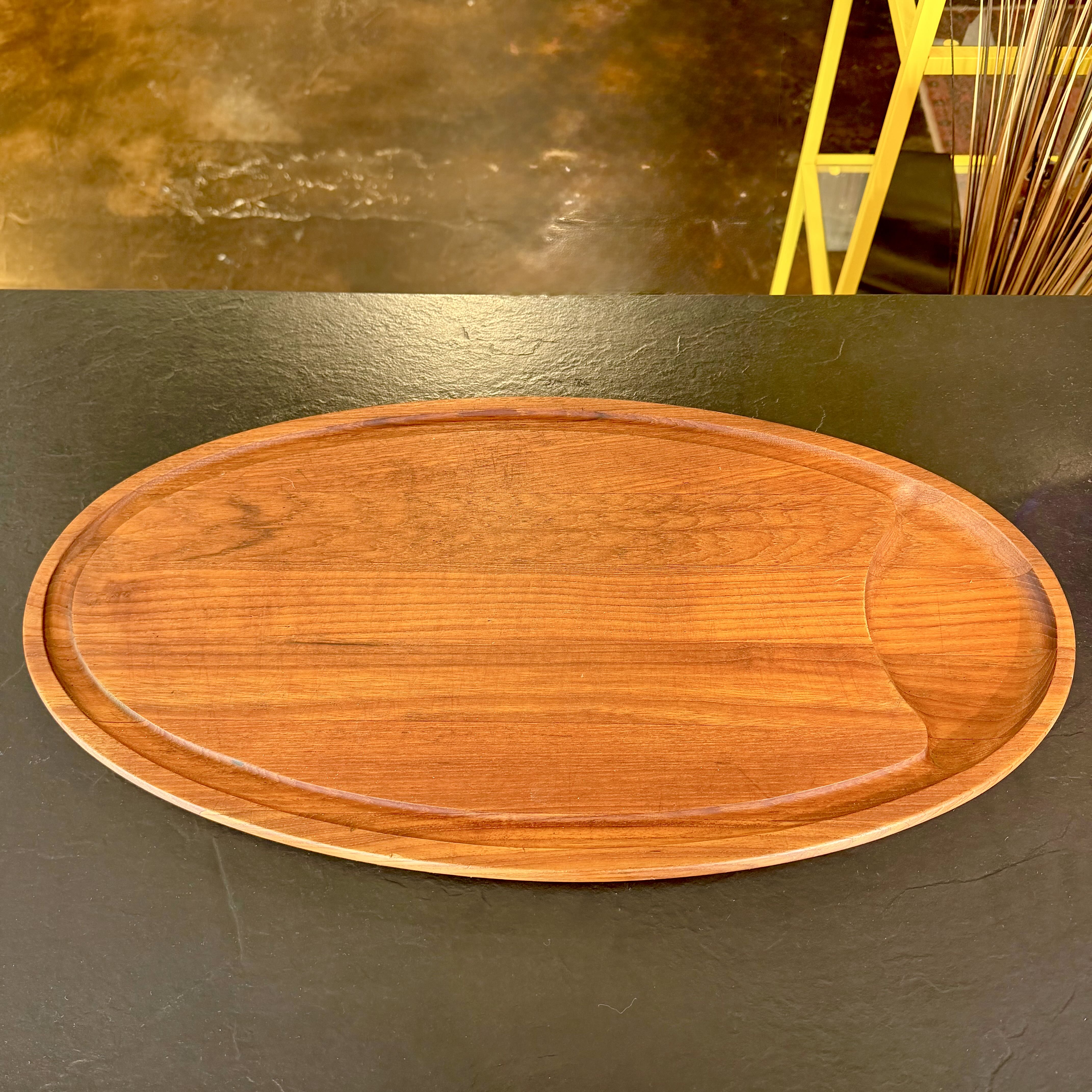 An oval wooden serving tray with a raised rim and a recessed center, shown on a dark tabletop with a blurred indoor background.