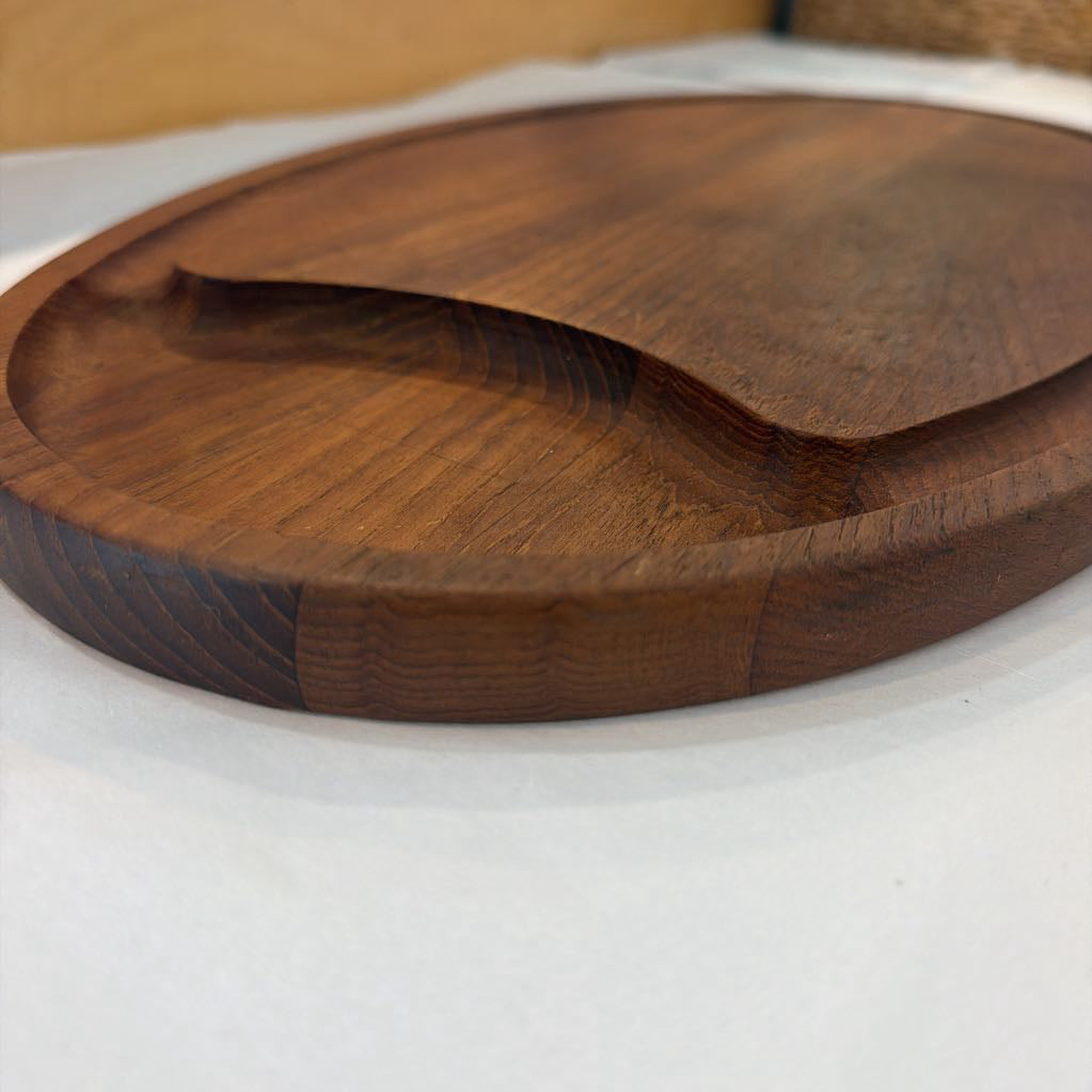 A round wooden serving board with a recessed center and a wavy cutout detail, shown on a white surface with a warm-toned background.
