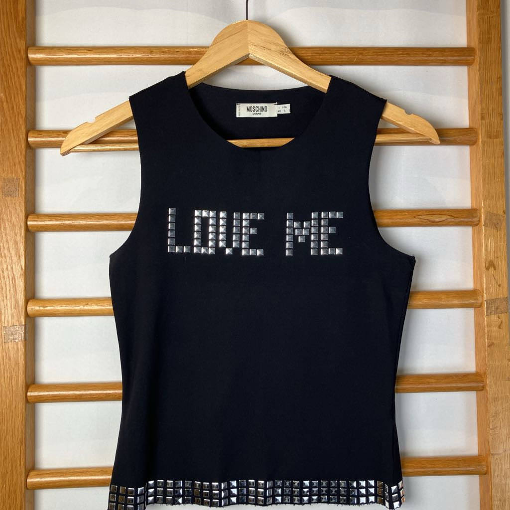Black sleeveless top with 'LOVE ME' text on a wooden hanger against a wooden lattice background