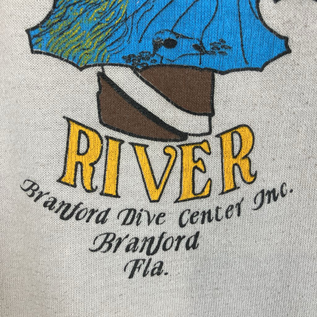 Close-up of a vintage-style printed graphic on a light beige fabric, featuring a blue and green fish illustration above the large yellow text 'RIVER' and smaller black text reading 'Branford Dive Center Inc. Branford Fla.