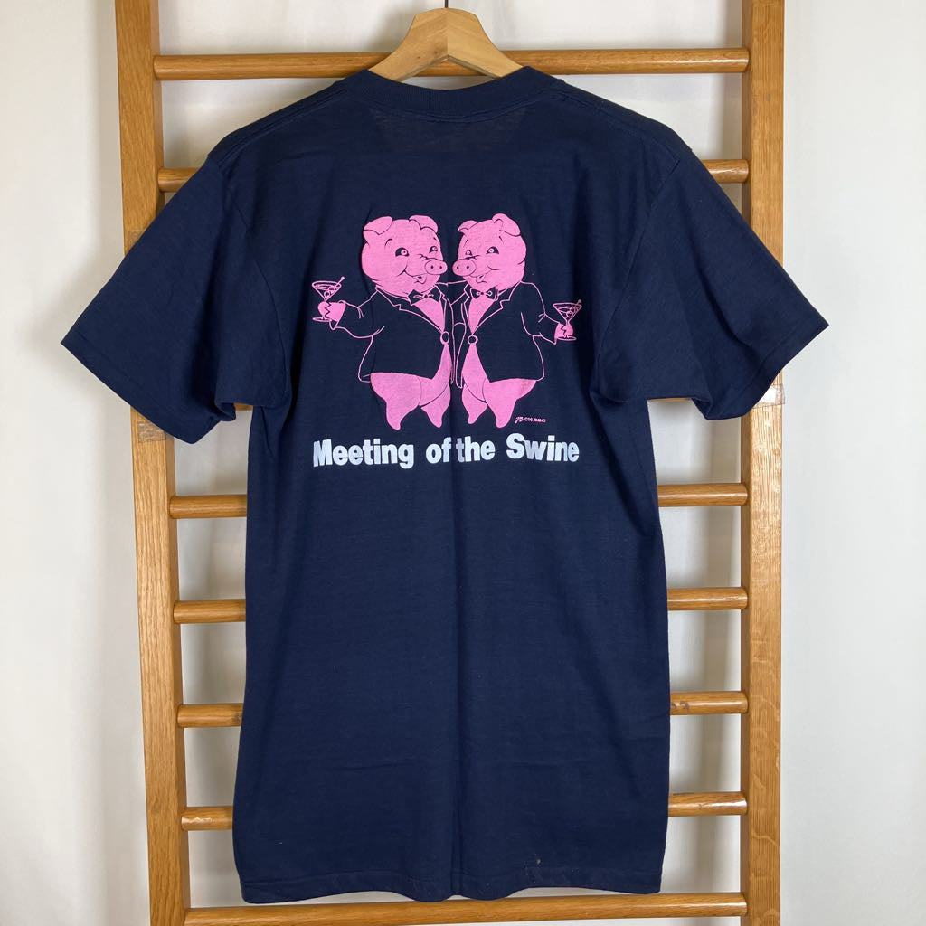 A navy blue t-shirt hanging on a wooden hanger against a wooden ladder. The back of the shirt features a pink graphic of two pig characters in suits holding drinks, with the white text 'Meeting of the Swine' underneath.