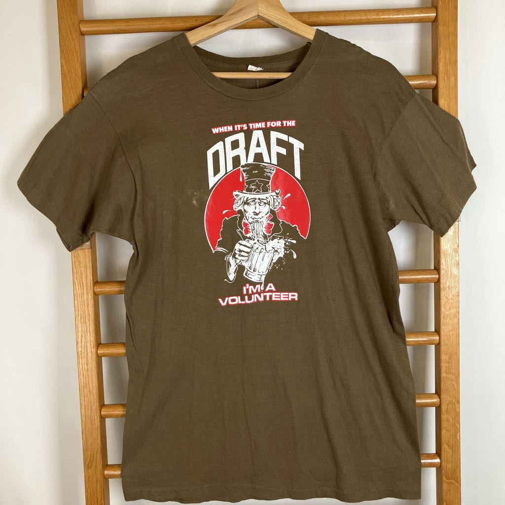 An olive brown short-sleeve graphic t-shirt hanging on a wooden hanger from a wooden ladder. The graphic features Uncle Sam with a red hat and a red circle background, with the text 'WHEN IT'S TIME FOR THE DRAFT I'M A VOLUNTEER'.