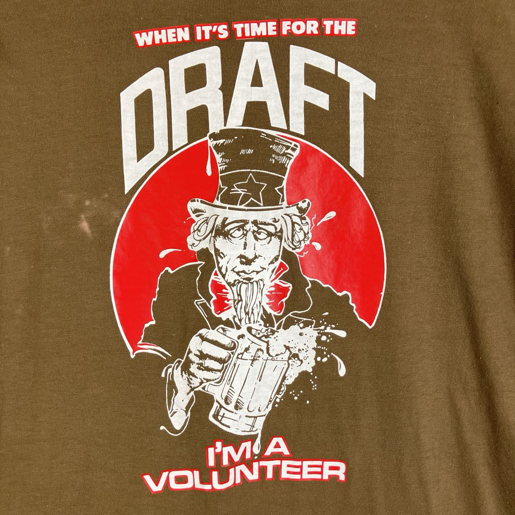 Close-up of a brown graphic t-shirt featuring a red, white, and black illustration of Uncle Sam drinking beer, with the text 'WHEN IT'S TIME FOR THE DRAFT' at the top and 'I'M A VOLUNTEER' at the bottom.