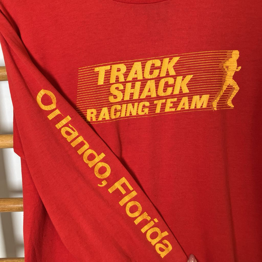 Red t-shirt with 'Track Shack Racing Team Orlando, Florida' text and graphic on a wooden surface.