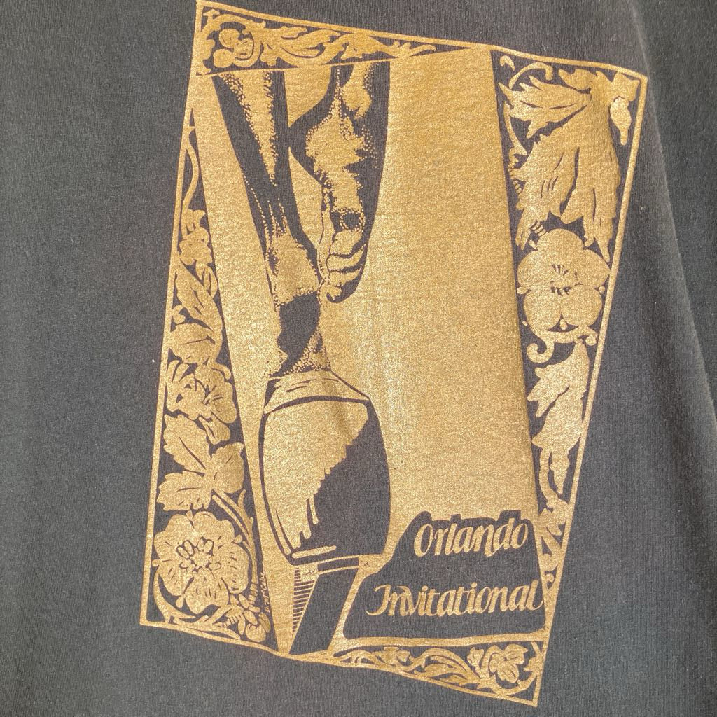 A close-up of a dark charcoal gray t-shirt featuring a vintage-style gold graphic print for the 'Orlando Invitational'. The graphic depicts a trophy with a severed foot and hand, framed by ornate floral borders, with the text 'Orlando Invitational' in the bottom right corner.